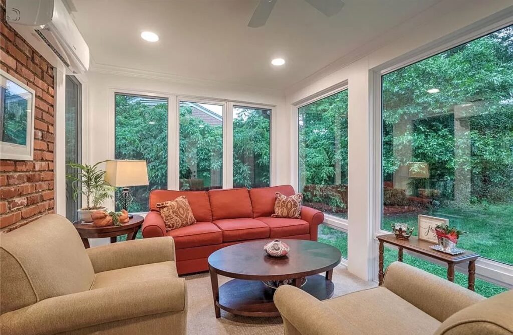 Living room with large windows showing green outdoor view, red sofa, beige armchairs, round coffee table, and exposed brick wall