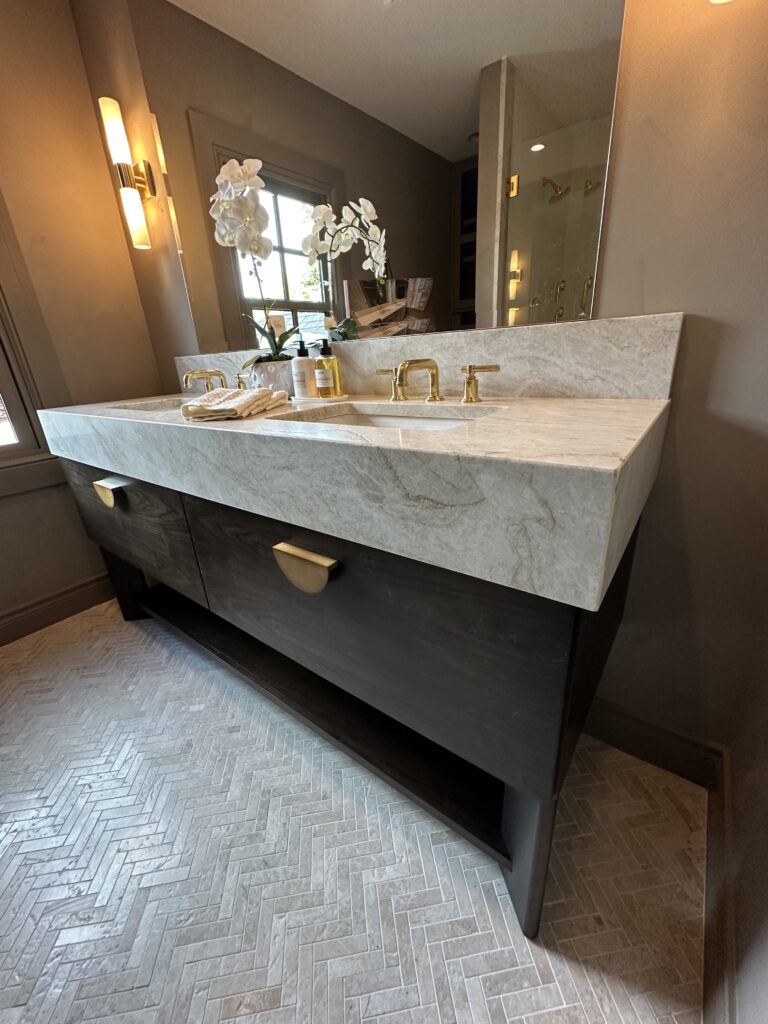 Modern bathroom with marble countertop, gold fixtures, dark wood vanity, herringbone tile floor, and orchid by window
