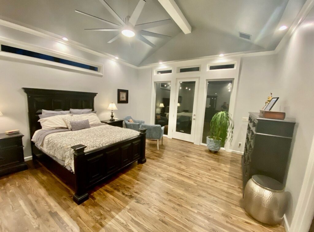 Large master bedroom with dark wood bed, hardwood floors, ceiling fan, and seating area near glass doors