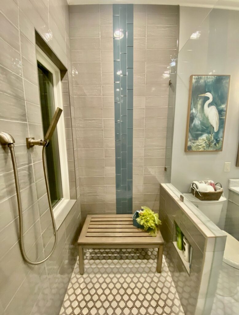 Shower area with a wooden slatted bench, gray and blue subway tiles, handheld shower head, and patterned floor tiles